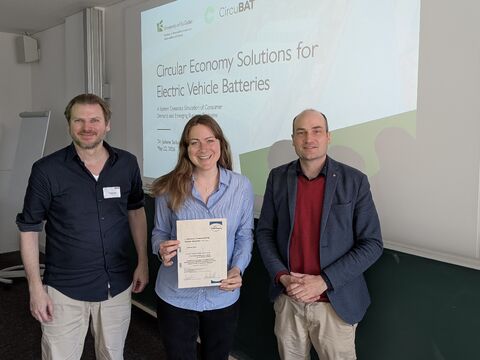 Three adults stand in a classroom like room the woman in the center holds a certificate with a projector showing Circular Economy Solutions for Electric Vehicle Batteries in the background